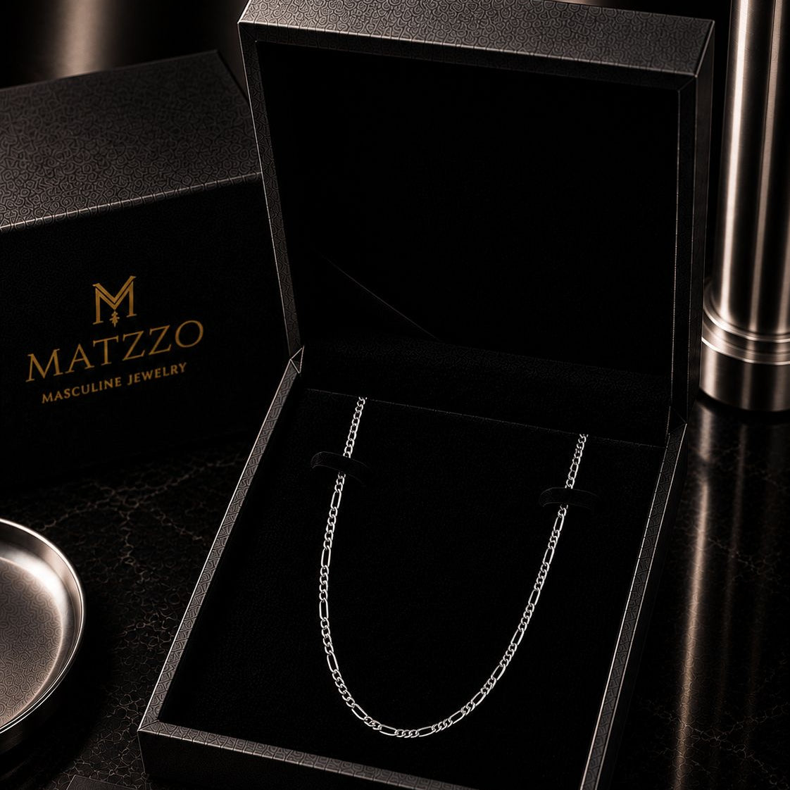 A silver-toned chain necklace displayed inside an open black MATZZO jewelry box, with matching branded boxes and a metallic cylindrical object on a dark surface.