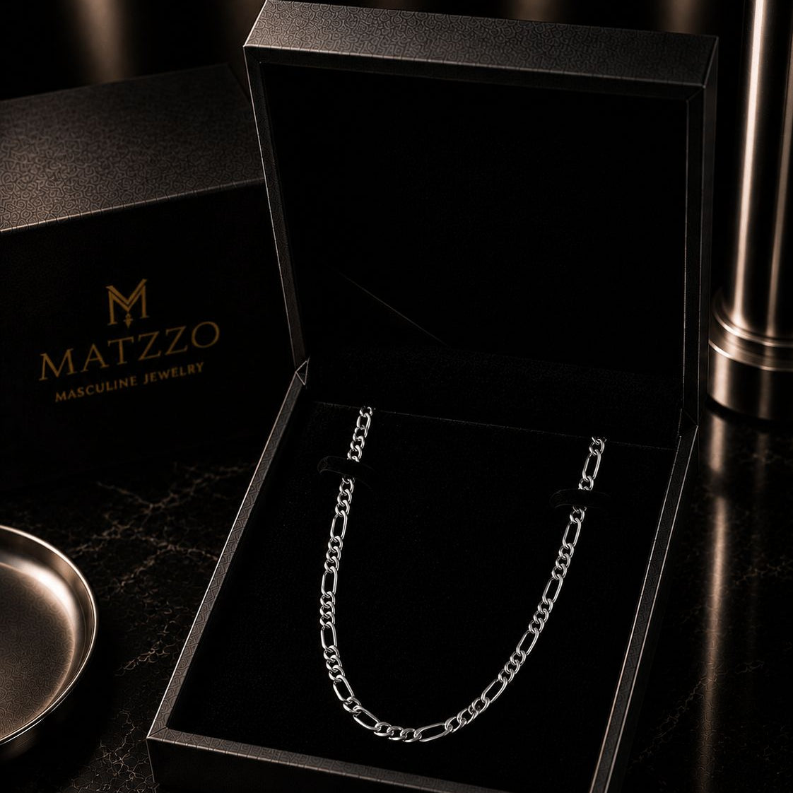 A silver Figaro chain necklace displayed inside an open black MATZZO jewelry box, with the brand's black packaging and card visible on a dark marble surface.